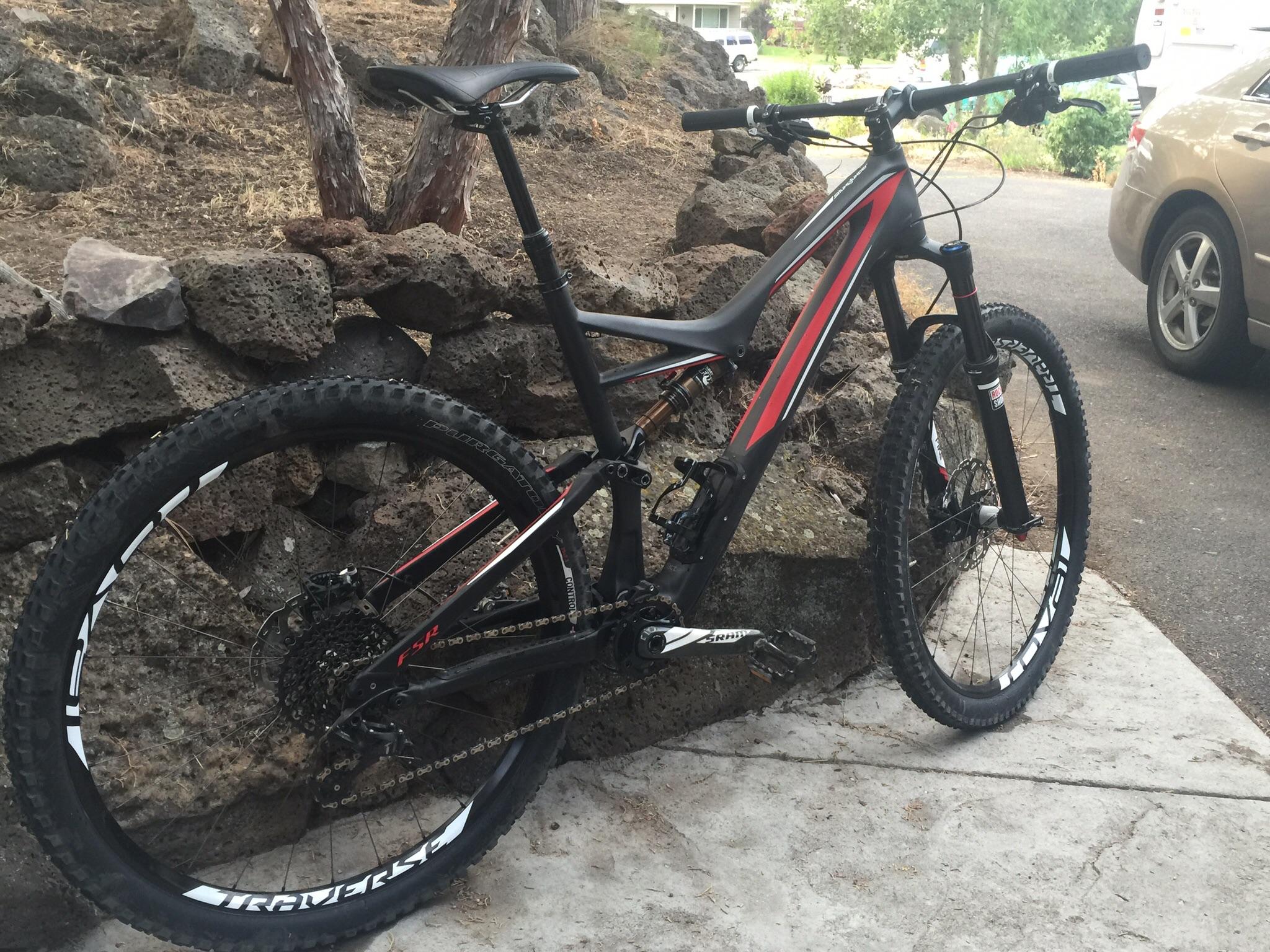 Specialized Stumpjumper FSR Pro Carbon: A mountain bike with a black and red frame is parked beside a stone wall. The bike features large, rugged tires and a visible chain system, showcasing its design for off-road riding. In the background, there is a gravel path and some greenery, hinting at a natural outdoor setting.