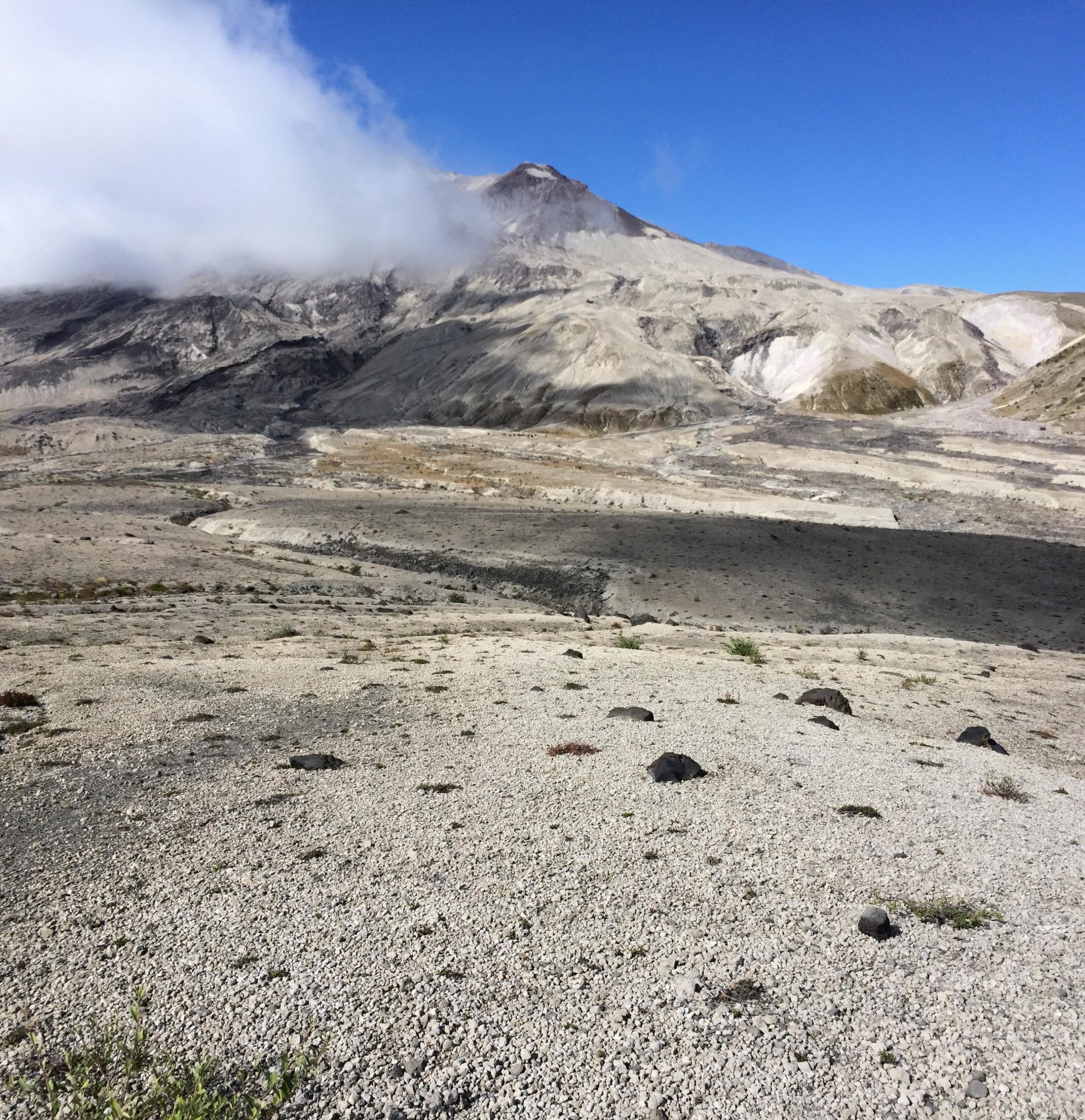 A barren landscape with gray volcanic rock and gravel leading to a mountain. The mountain is partially shrouded in clouds under a clear blue sky. The scene showcases the aftermath of a volcanic eruption, with minimal vegetation and a rugged terrain. Ape Canyon#234, Abraham#216d, Smith Creek#225 Trails mountain bike trail.