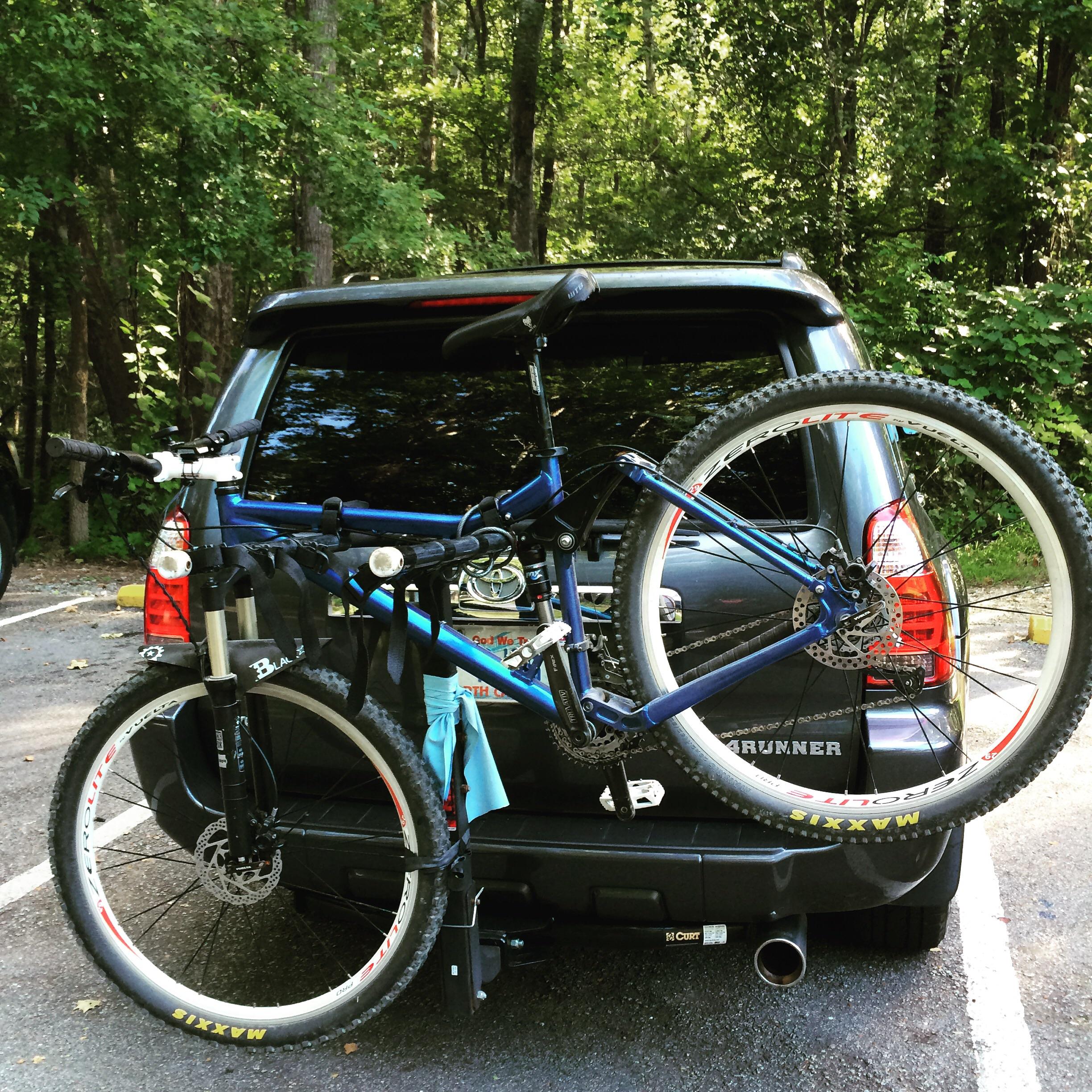 A blue mountain bike is securely mounted on the back of a black SUV parked in a wooded area. The bike is attached to a bike rack, with the SUV's tailgate visible in the background, along with greenery surrounding the vehicle. Enterprise South mountain bike trail.