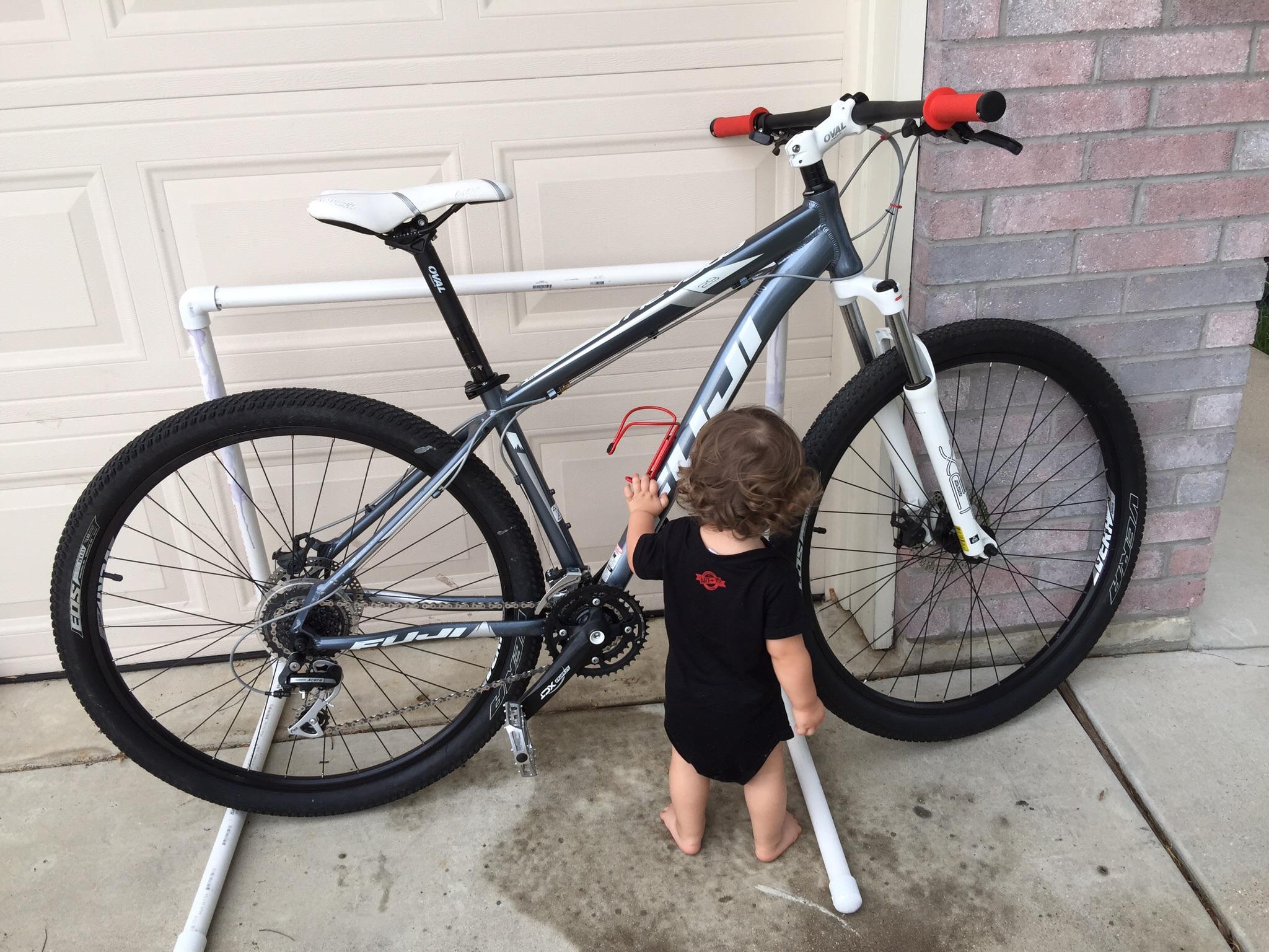Fuji Nevada 29 1.7: A young child stands next to a mountain bike that is secured on a bike stand in a driveway. The bike features a gray and white frame with red handlebars, and the child is curiously interacting with the bike. The background includes a garage door and a brick wall.