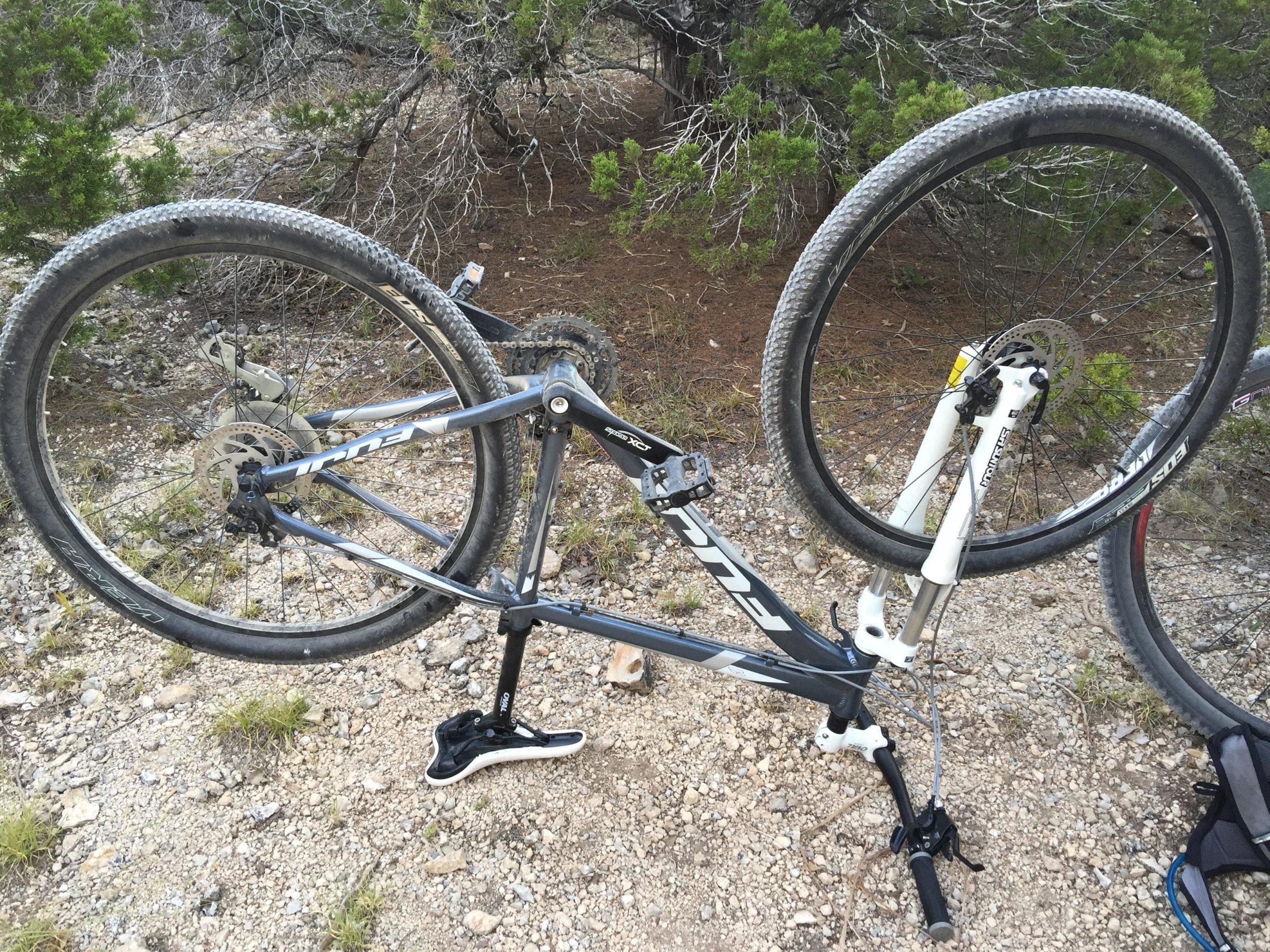 A mountain bike positioned upside down on a gravel surface, with its front wheel elevated and the rear wheel resting on the ground. The bike features a dark frame with branding, and both wheels have knobby tires. Surrounding vegetation includes low shrubs and trees in the background. Dana Peak mountain bike trail.