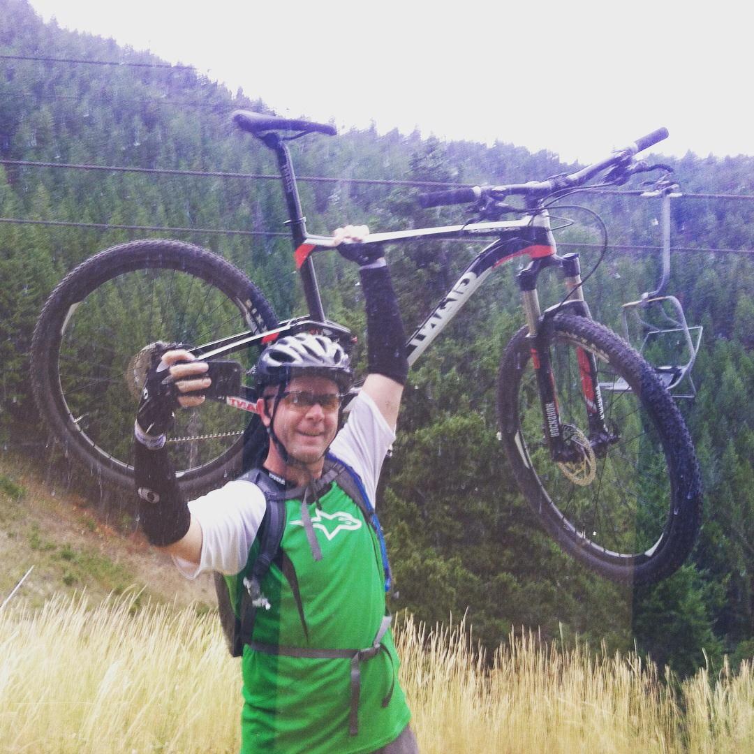 A mountain biker wearing a green jersey and helmet poses with a bicycle lifted above his head, smiling triumphantly amidst a scenic backdrop of trees and grass. The image captures the joy of biking and the thrill of outdoor adventure. Bald Mountain Bike Park mountain bike trail.