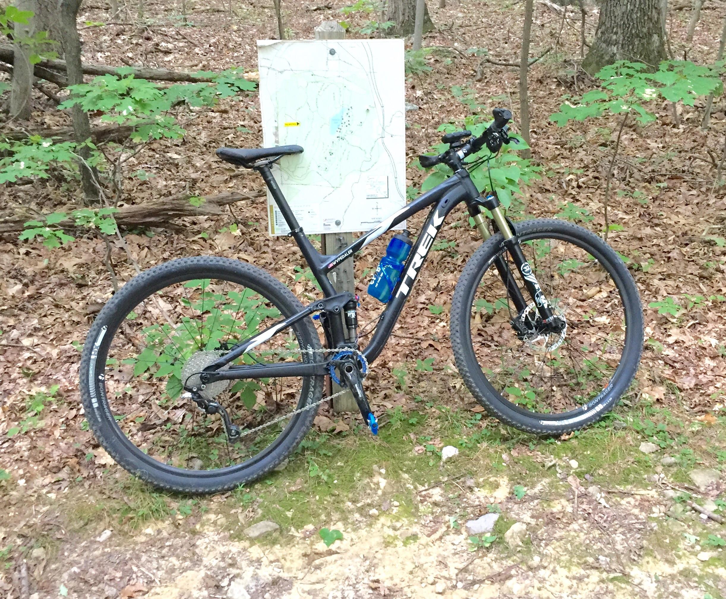 A mountain bike leaning against a trail map sign in a wooded area, surrounded by green leaves and fallen brown foliage. The bike features a sturdy frame, thick tires, and a water bottle attached to its frame. The map in the background provides information about the trails in the vicinity. Greenbrier State Park mountain bike trail.