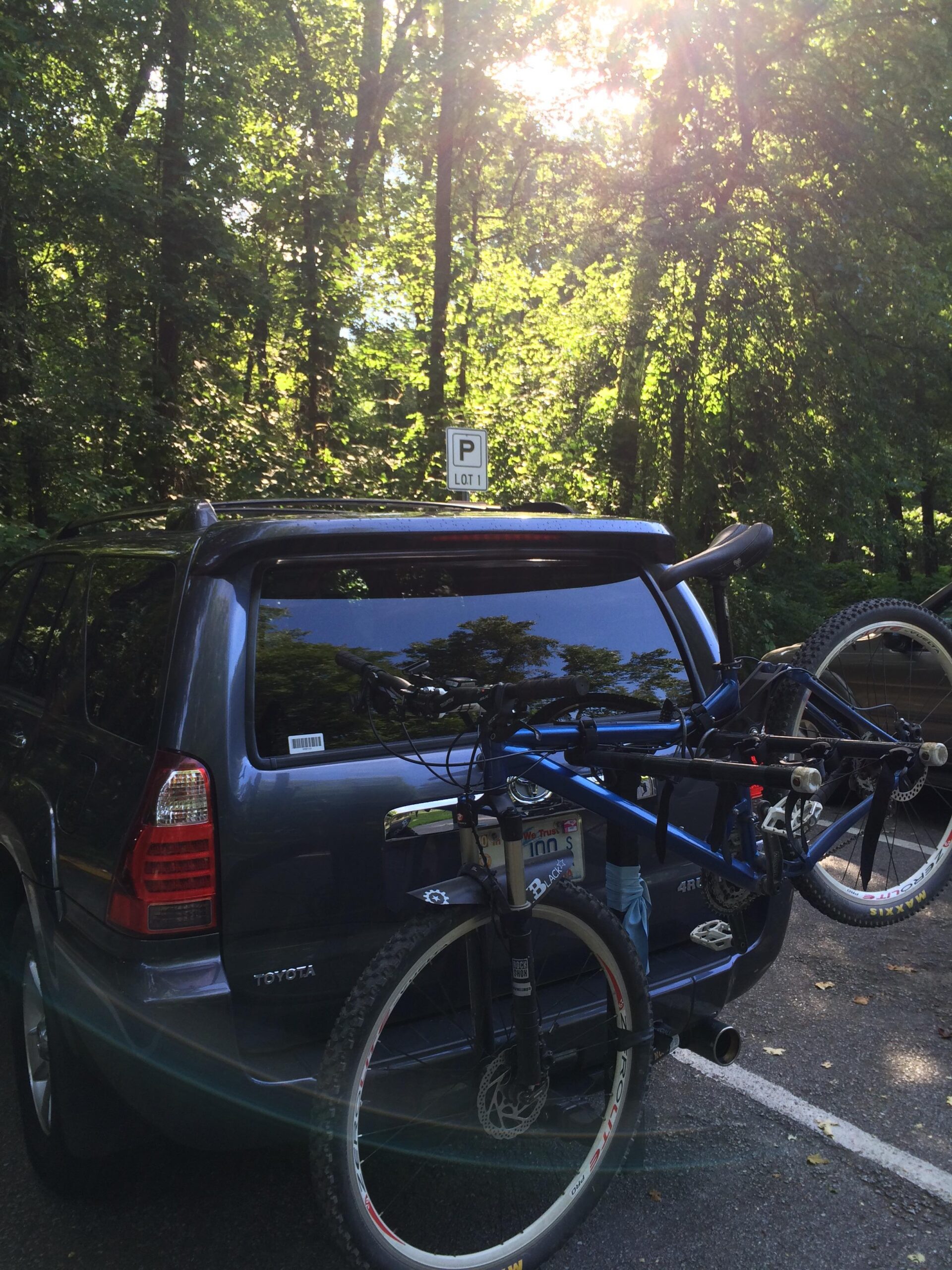 A blue Toyota SUV parked in a wooded area, with a mountain bike secured to the back using a bike rack. Sunlight filters through the trees, creating a warm, natural ambiance. A parking sign labeled "P Lot 1" is visible in the background. Enterprise South mountain bike trail.
