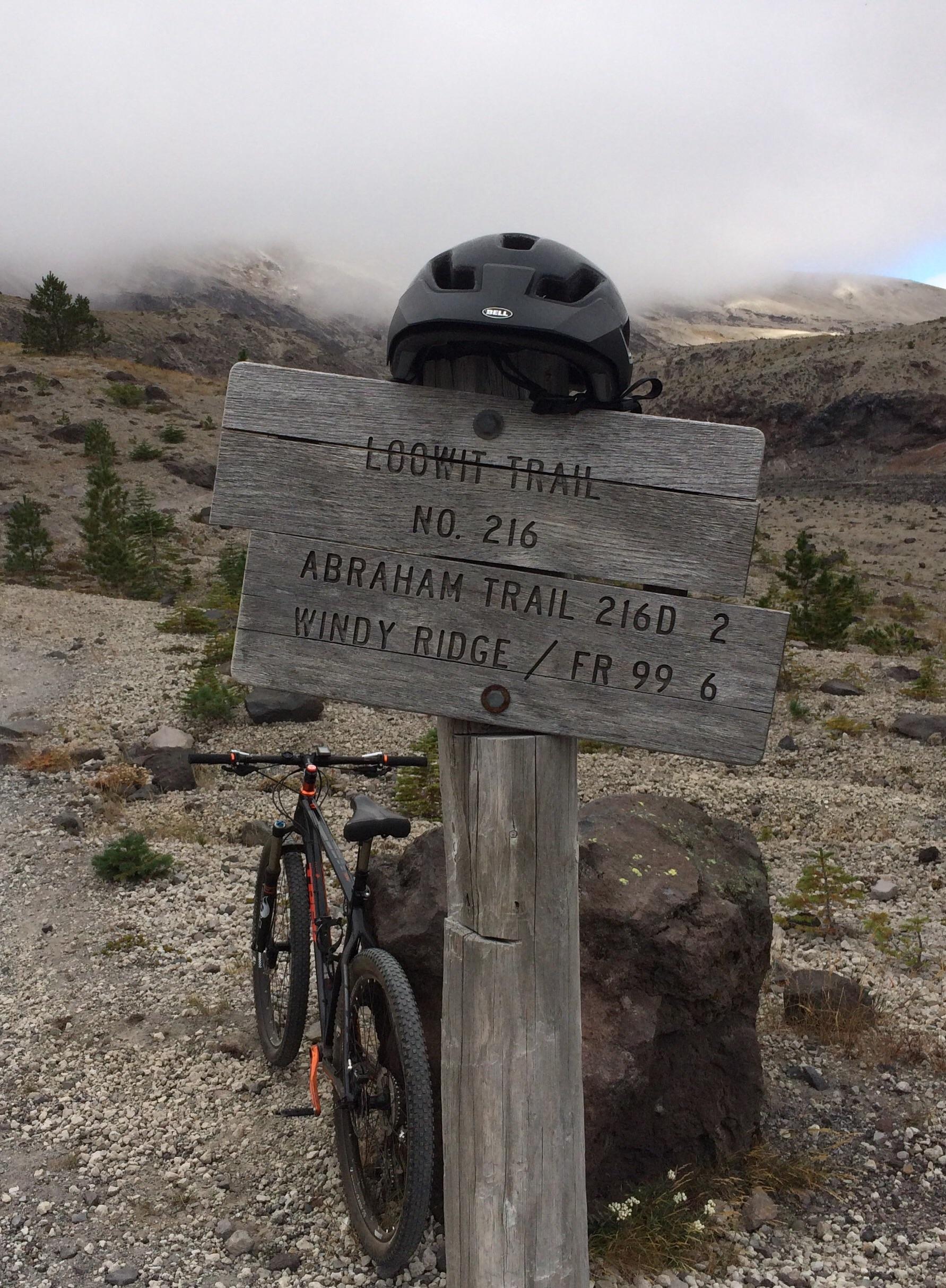 A wooden trail sign displaying directions for the Loowit Trail (No. 216), Abraham Trail (216D), and Windy Ridge (FR 99) surrounded by a rocky landscape. A mountain bike is parked beside the sign, and a black helmet rests on top of it. Fog covers the mountains in the background, creating a misty atmosphere. Ape Canyon#234, Abraham#216d, Smith Creek#225 Trails mountain bike trail.