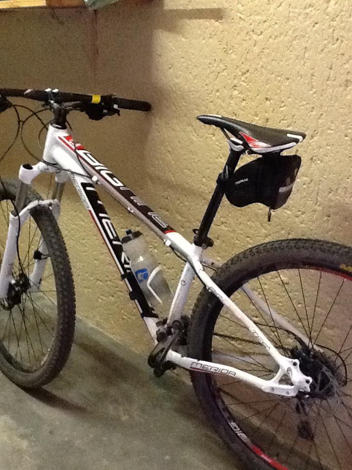 Merida Big.Nine 900: A white mountain bike with red and black accents is parked against a textured wall. It features a water bottle holder, a black saddle, and thick, knobby tires suited for off-road terrain. The bike is positioned in a garage or storage space with dim lighting and a simple background.