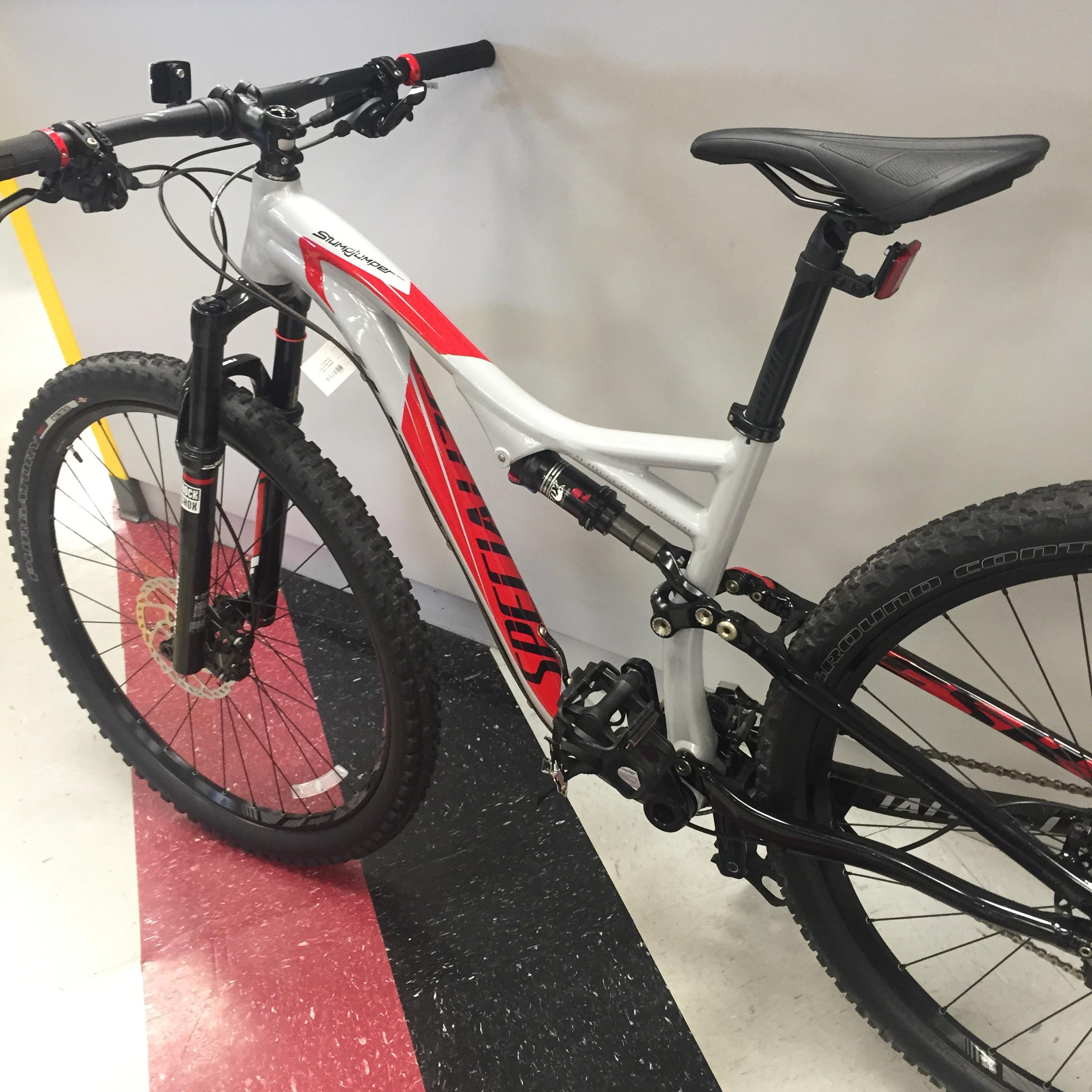Specialized Stumpjumper FSR: A modern mountain bike with a gray and red frame, featuring wide tires, front suspension, and a sleek design, leaning against a wall in a store. The floor has a black and red pattern, adding to the store's aesthetic.