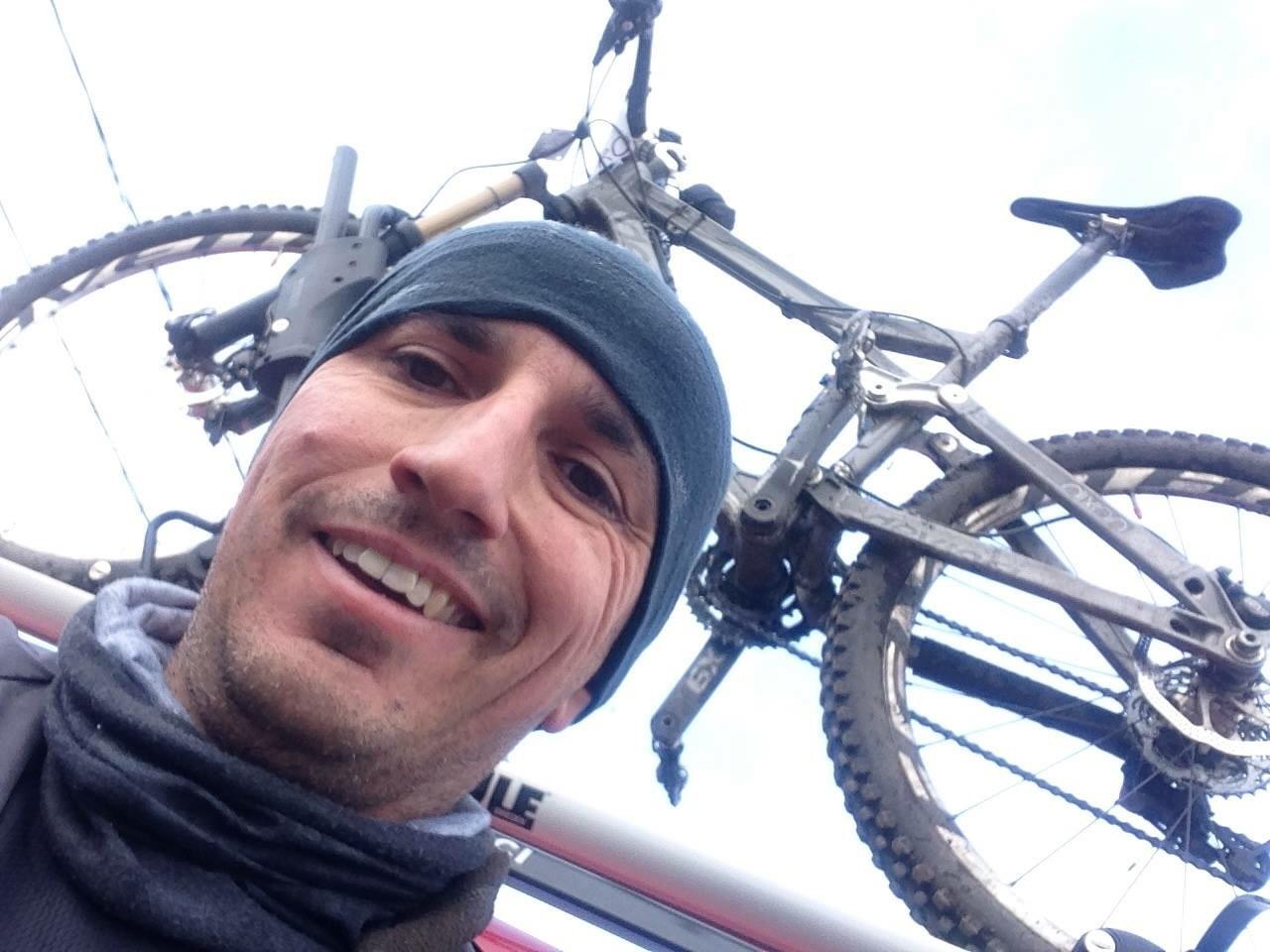 Devinci Dixon RS: A man wearing a beanie and scarf smiles for a selfie with a mountain bike mounted above him, set against a cloudy sky.