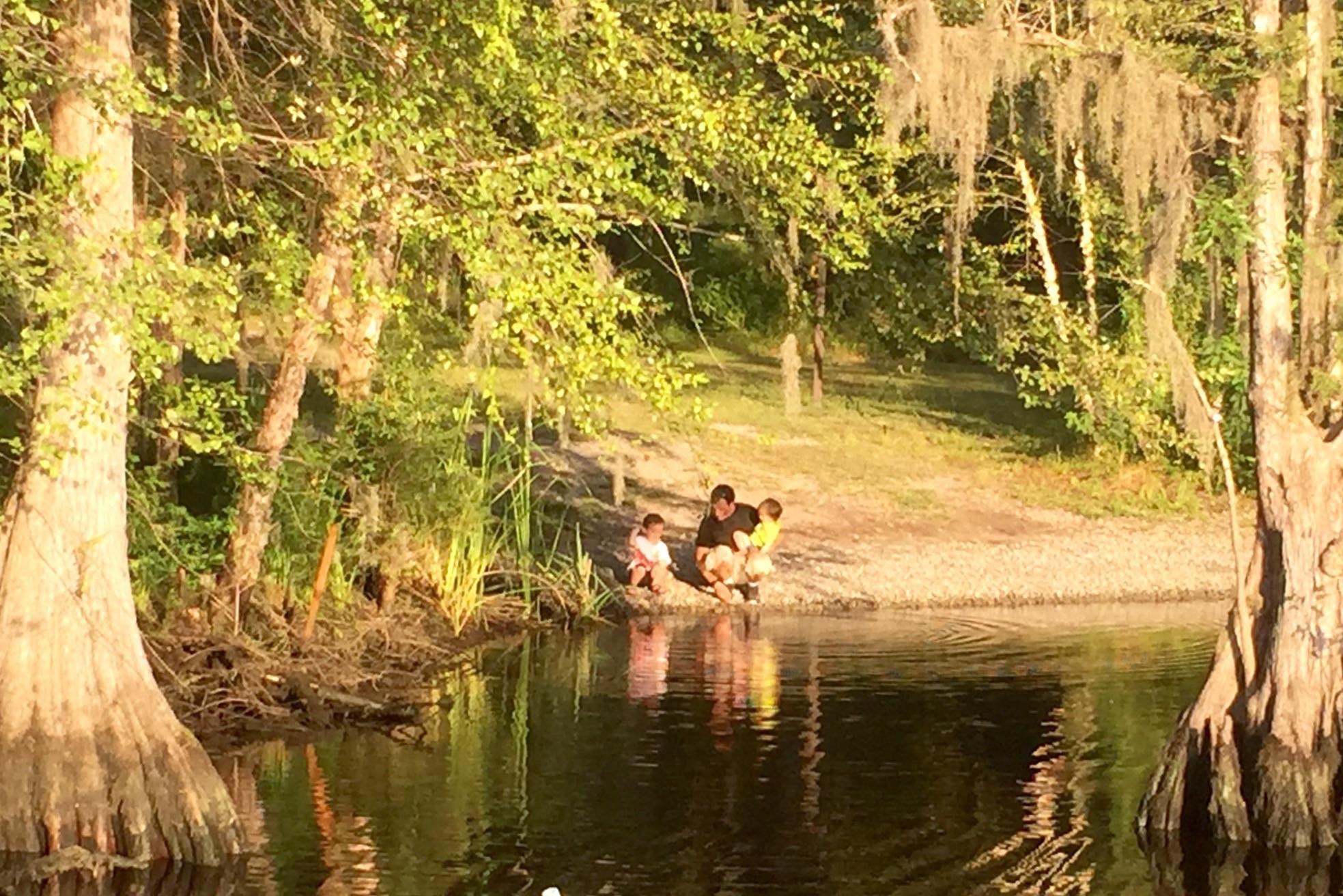 Three children sit by the edge of a calm body of water, playing and looking into the water. Surrounded by lush greenery and trees with Spanish moss, the scene captures a peaceful moment in nature during golden hour, with sunlight filtering through the leaves. Jackson Bluff Trail System mountain bike trail.