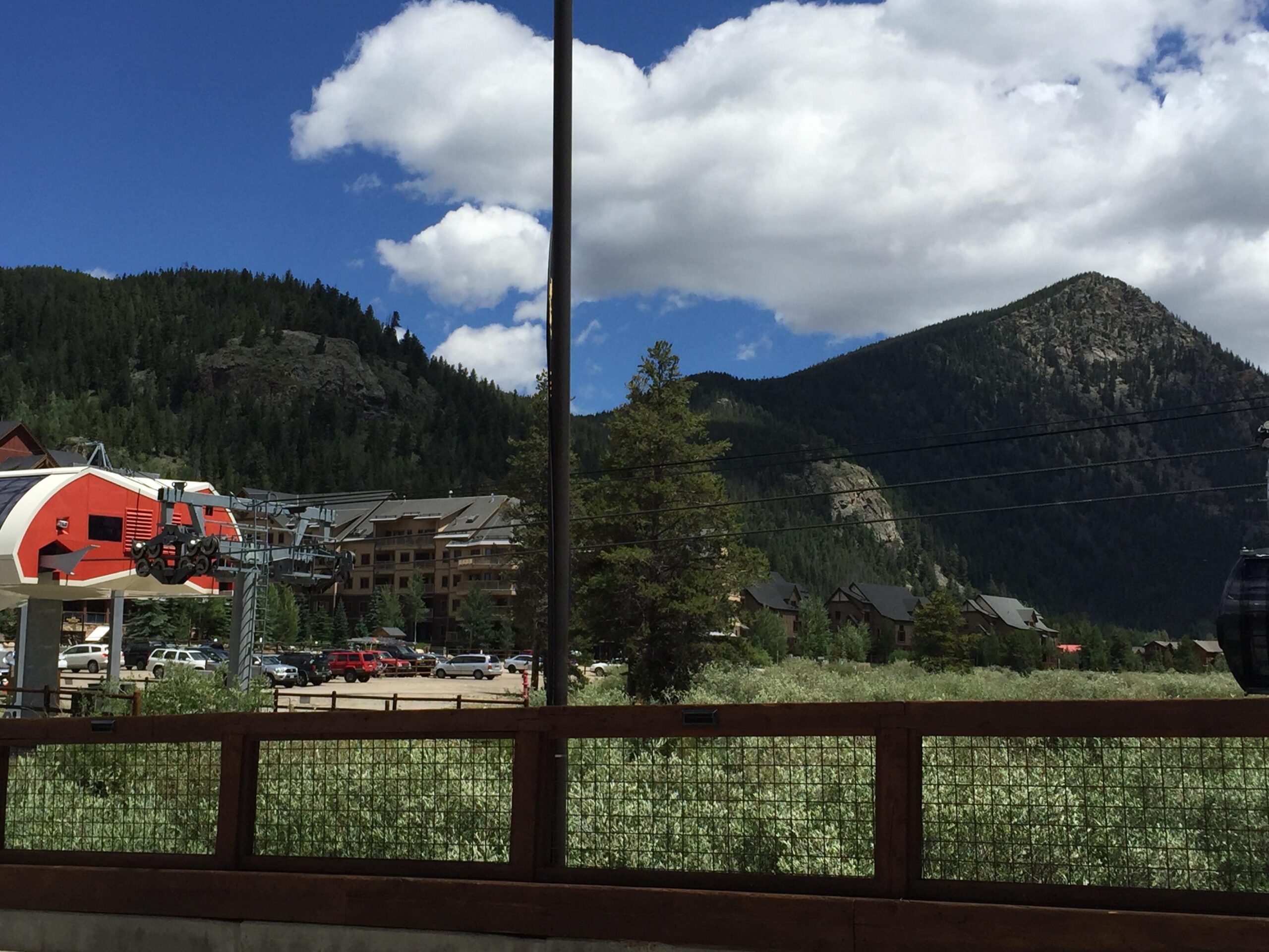 A scenic view featuring a ski lift station with red and white gondolas, set against a backdrop of lush green mountains and a bright blue sky with fluffy white clouds. In the foreground, there is a wooden fence, and surrounding the lift station are several buildings and parked cars, indicating a mountain resort area. Keystone Resort Bike Park mountain bike trail.