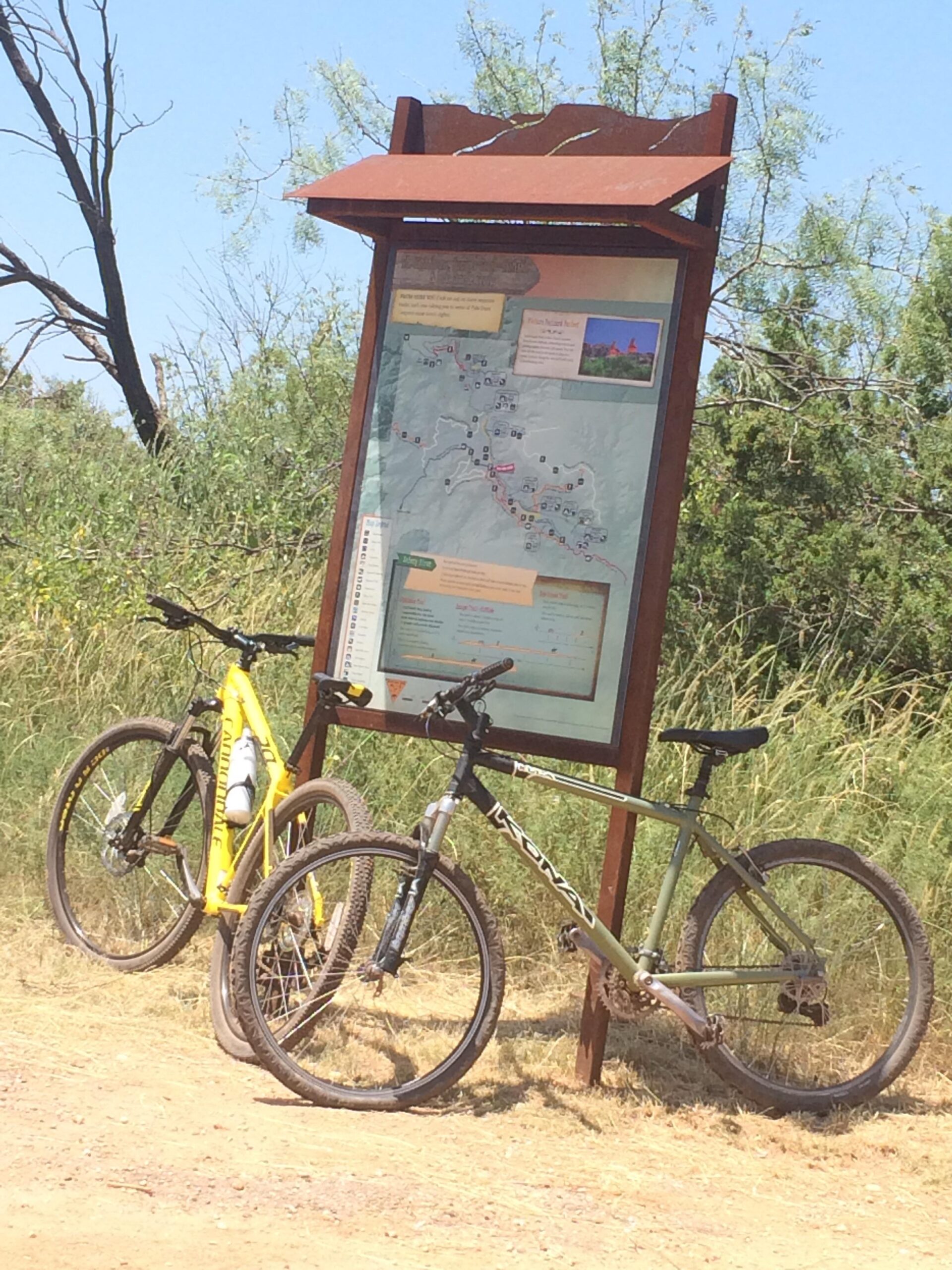 Kona Kahuna: Two mountain bikes are parked beside a trail map sign in a grassy area. The sign provides information about nearby trails, with various marked routes visible on the map. The scene is set in a sunny environment with clear blue skies.
