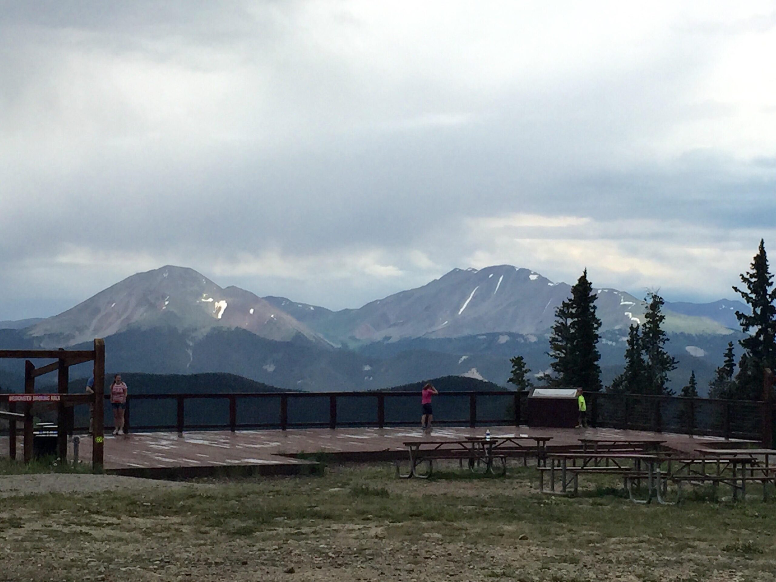 A panoramic view of distant mountains under a cloudy sky, with a wooden deck in the foreground. Two individuals stand on the deck, while picnic tables are visible nearby. The landscape features pine trees and patches of snow on the mountain peaks. Keystone Resort Bike Park mountain bike trail.