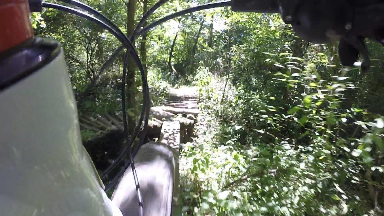 A close-up perspective from the handlebars of a vehicle navigating a narrow, wooded trail. Lush green foliage surrounds the path, with sunlight filtering through the trees. A section of the trail is visible ahead, featuring wooden planks and a rustic terrain. Rangeline Nature Preserve mountain bike trail.
