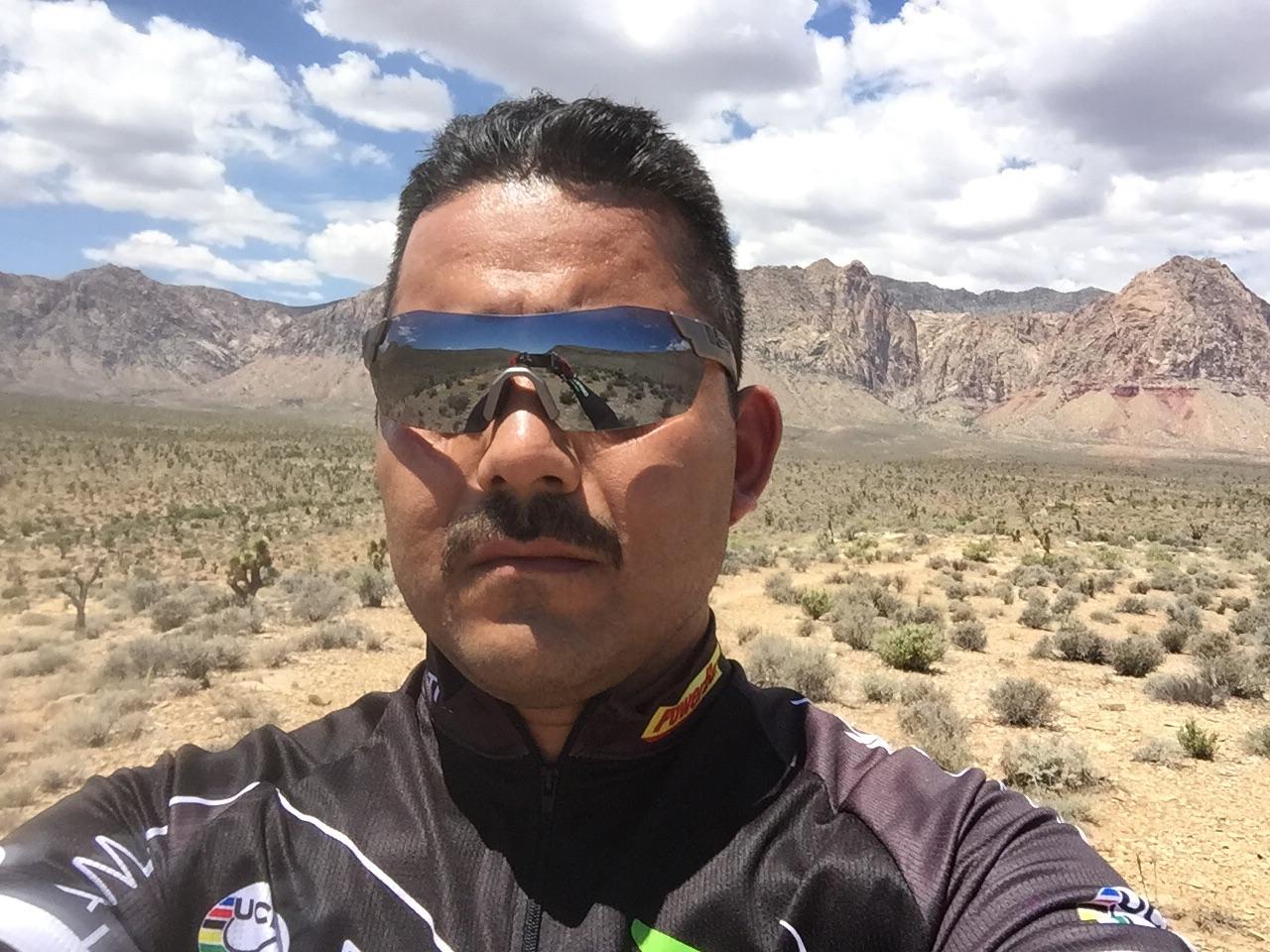 A cyclist taking a selfie in a desert landscape with mountains in the background, wearing sunglasses and a cycling jersey, under a partly cloudy sky. Blue Diamond mountain bike trail.