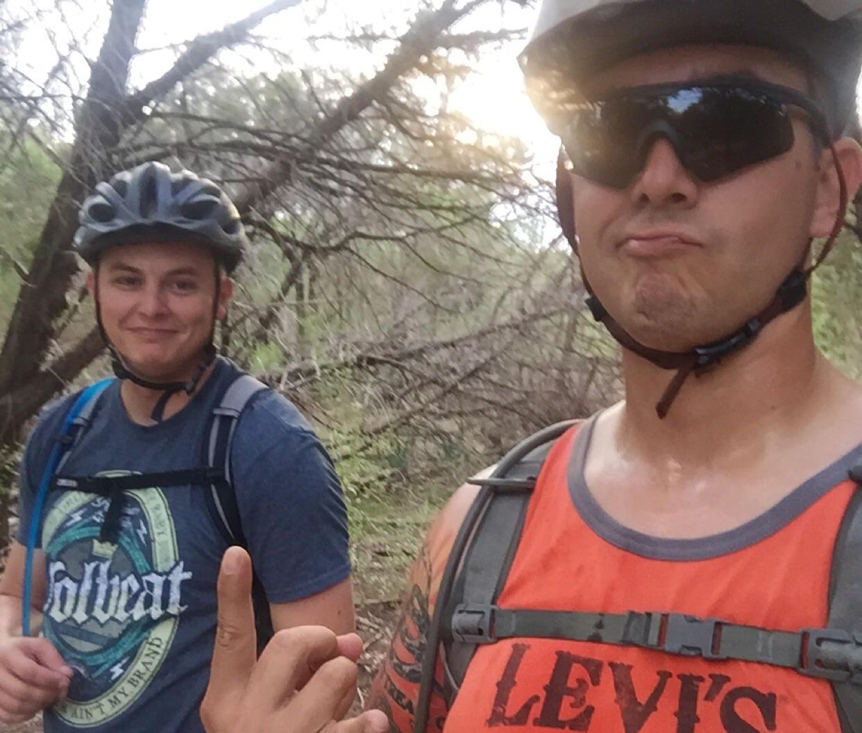 Two individuals wearing helmets and sunglasses are posing for a selfie while mountain biking on a wooded trail. One person is making a playful gesture with their hand, while the other is smiling in the background. The setting includes trees and natural foliage, suggesting an outdoor adventure. Dana Peak mountain bike trail.