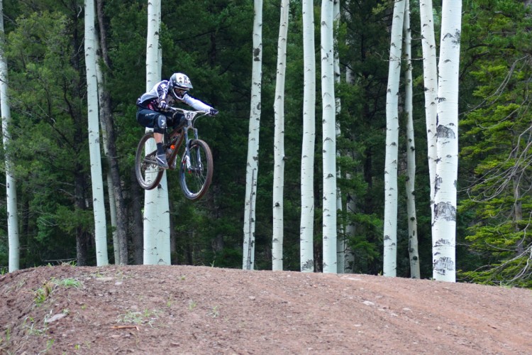 A mountain biker in mid-air performing a jump over a dirt mound, surrounded by tall white aspen trees and lush green foliage in the background.