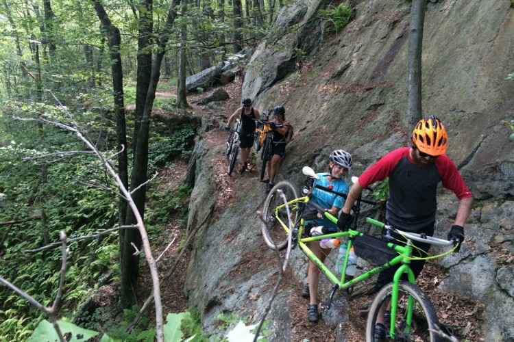 Four mountain bikers navigating a rocky trail in a wooded area, with two riders carrying their bikes while another two maneuver along the path. The scene features lush green foliage, trees, and rugged terrain.