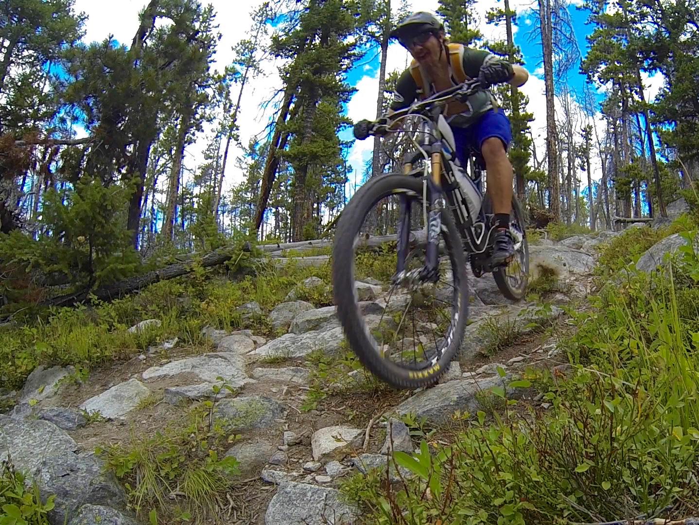 A mountain biker navigating a rocky trail in a forested area, surrounded by tall trees and vibrant greenery, under a partly cloudy blue sky. The biker is captured mid-motion, showcasing an athletic posture as they maneuver over the uneven terrain. MacDonald Pass mountain bike trail.