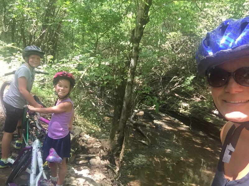 A woman and two children posing beside their bicycles near a creek, surrounded by lush greenery. The kids are wearing helmets and smiling, enjoying a sunny day outdoors. Knucklehead mountain bike trail.
