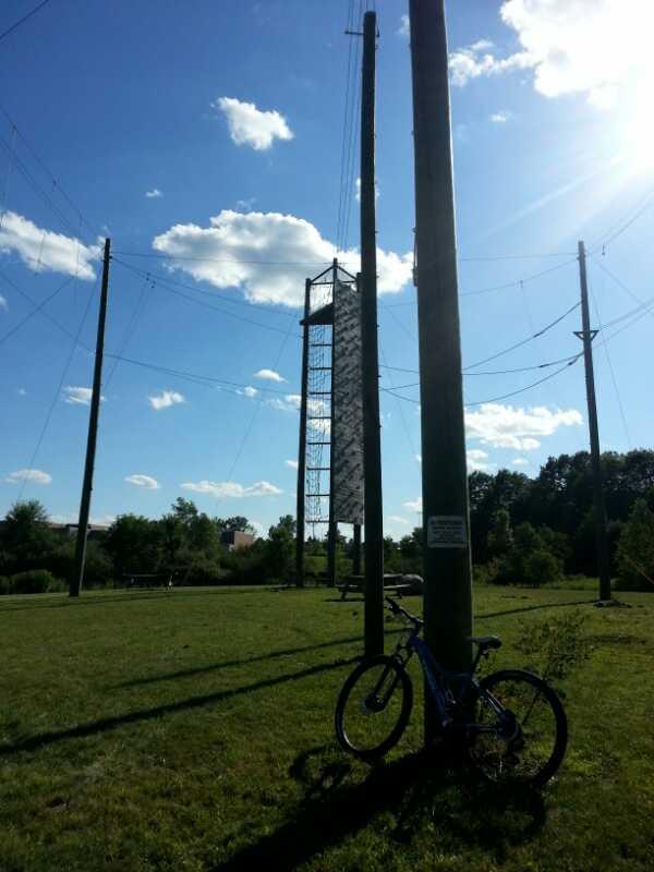 A clear blue sky with scattered clouds above a grassy field. In the foreground, a blue bicycle is leaning against a utility pole. In the background, a tall structure with a climbing wall is visible, surrounded by additional utility poles and power lines. Trees line the edge of the field, creating a natural backdrop. Lake Orion High School mountain bike trail.