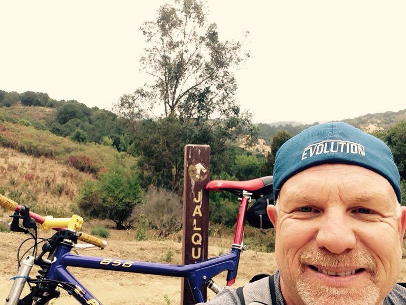 A man smiling for a selfie in front of a hiking trail sign that reads "JALQU." A colorful mountain bike is parked beside him, and the background features a hilly landscape with greenery and overcast skies. Anthony Chabot Regional Park mountain bike trail.