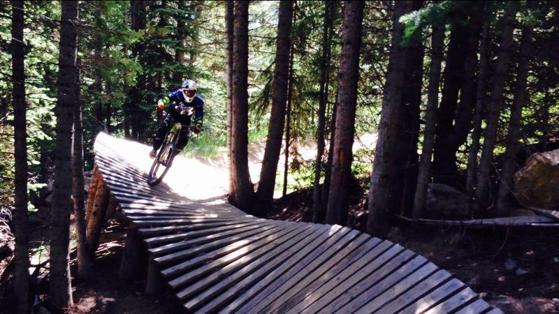A mountain biker riding on a wooden beam pathway through a dense forest, surrounded by tall trees and dappled sunlight. Trestle Bike Park mountain bike trail.