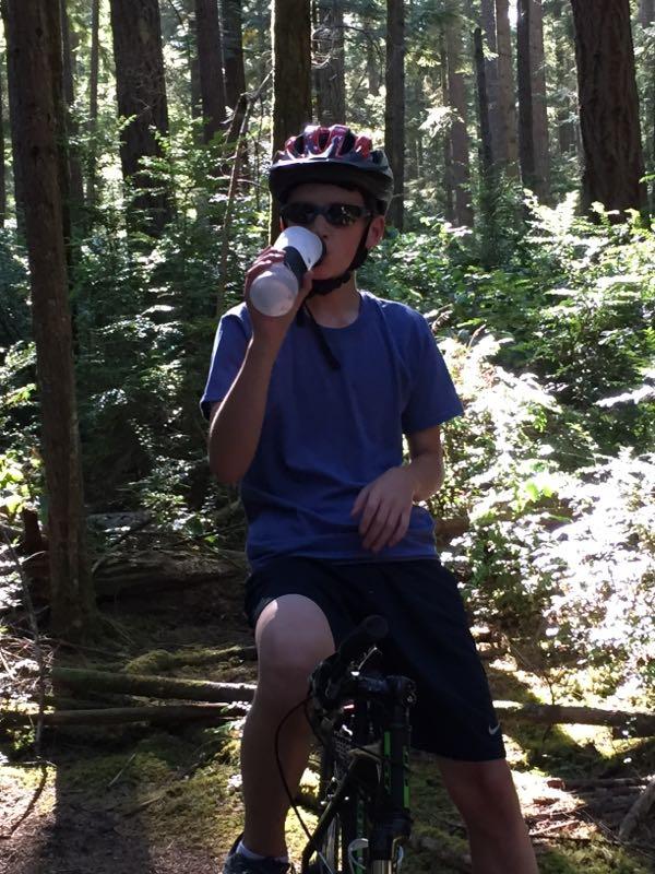 A young person wearing sunglasses and a helmet sits on a mountain bike in a forested area, sipping from a cup. Sunlight filters through the trees, illuminating the greenery around them. Banner Forest mountain bike trail.