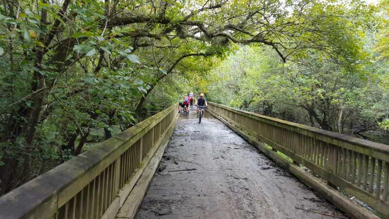 A wooden bridge surrounded by lush greenery, with cyclists riding along a muddy path. The scene is framed by overhanging branches, creating a natural canopy. Little Big Econ State Forest mountain bike trail.