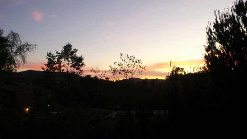 A serene sunset view with silhouettes of trees and hills against a colorful sky, featuring shades of orange, pink, and purple. The scene captures the tranquil beauty of nature during dusk. Lake Hodges mountain bike trail.