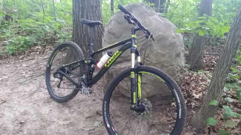 A mountain bike resting on a dirt trail surrounded by trees, with a large rock in the background. The bike features a black frame with green accents and is equipped with a water bottle attached to its frame. Leaves and foliage can be seen on the ground and in the surrounding area. Holdridge Recreation Area mountain bike trail.