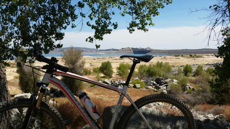 A mountain bike rests against a tree, with a view of a dry lakebed and distant hills. The scene is framed by green leaves, showcasing a sunny day with blue skies above. Granite Bay Trail mountain bike trail.