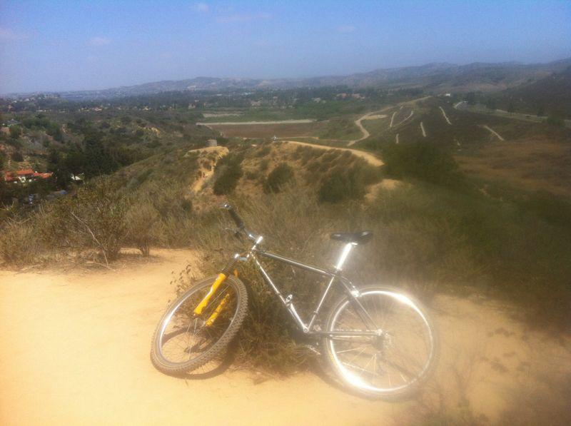 A mountain bike resting on a sandy trail overlooking a scenic landscape with rolling hills and sparse vegetation under a clear blue sky. In the background, a valley with houses can be seen. Peters Canyon Regional Park mountain bike trail.