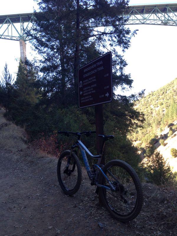 Mountain bike leaning against a trail sign at the intersection of Bridgeview Trail and Stagedoad Trail, with a view of a bridge and surrounding greenery in the background. Stagecoach / Flood / Manzanita mountain bike trail.