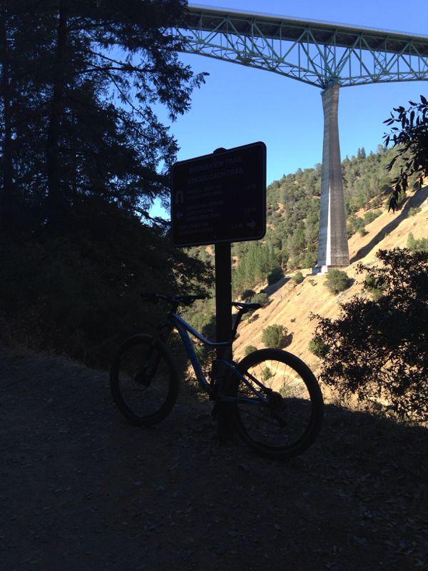 A mountain bike leaning against a trail sign with a bridge overhead. The scene is set in a forested area with hills in the background, showcasing a clear blue sky. Stagecoach / Flood / Manzanita mountain bike trail.