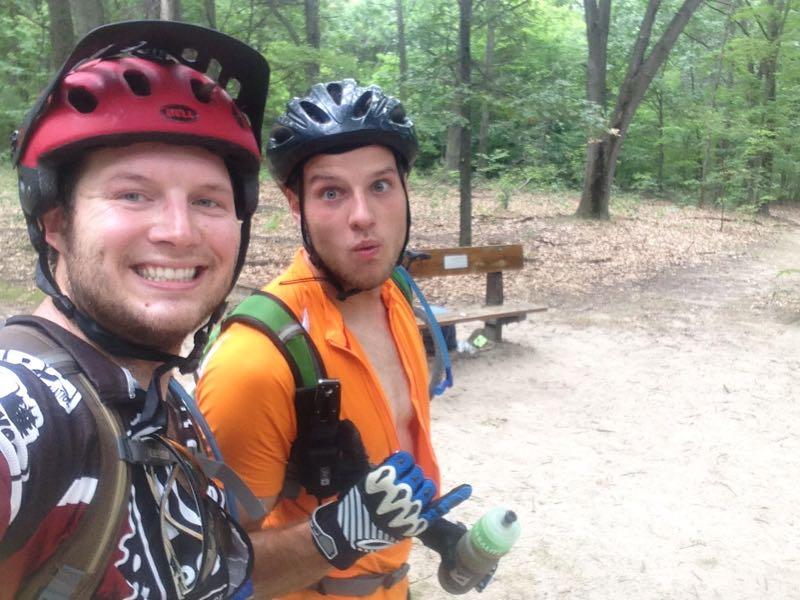 Two cyclists taking a selfie in a forested area. One is wearing a red helmet and a black biking jersey, smiling broadly, while the other, in an orange shirt, is making a surprised expression. They both have gloves on and are holding a water bottle. In the background, there's a wooden bench and trees surrounding them. Potawatomi trail mountain bike trail.