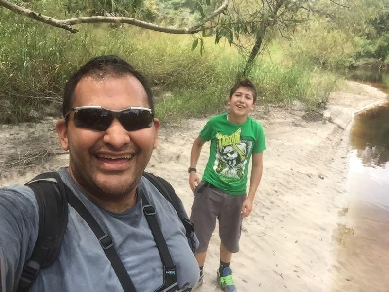 A smiling adult and a young boy pose for a selfie by a riverbank, surrounded by greenery. The adult is wearing sunglasses and a gray shirt with straps, while the boy is in a green t-shirt and shorts, looking cheerful. The background features sandy shore and lush foliage. Little Big Econ State Forest mountain bike trail.