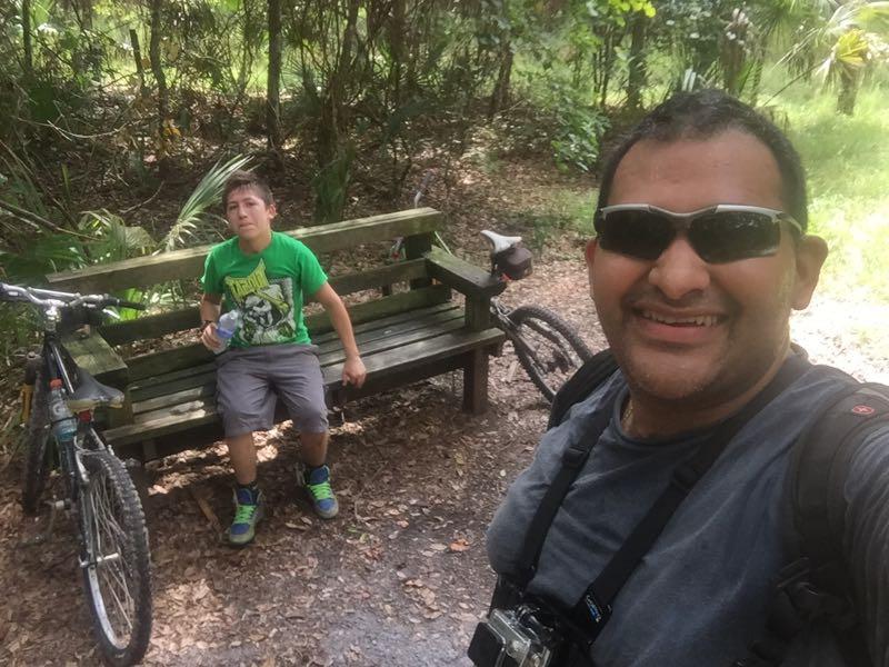 A man and a boy resting on a wooden bench in a wooded area, with two bicycles parked nearby. The boy is wearing a green t-shirt and shorts, holding a water bottle, and looking away from the camera. The man is smiling and taking a selfie, wearing sunglasses and a camera strap across his shoulder. The scene is surrounded by lush greenery. Little Big Econ State Forest mountain bike trail.