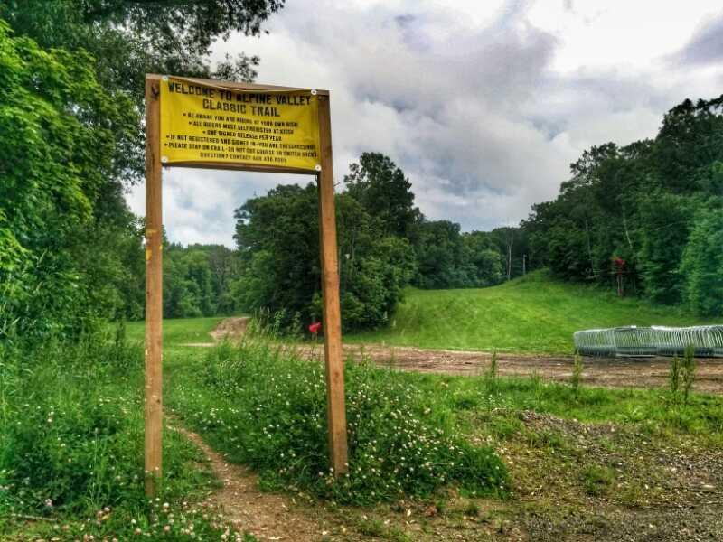 Sign at the entrance of the Alpine Valley Classic Trail, with guidelines and information for visitors, surrounded by greenery and a clear sky above. The trail path is visible in the background, leading towards a hilly area. Alpine Valley mountain bike trail.