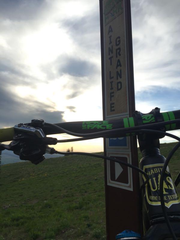 Close-up view of a mountain bike handlebar in front of a trail sign with the words "Ain't Life Grand." The background features a grassy landscape under a cloudy sky, suggesting a scenic outdoor setting. Peaked mountain bike trail.