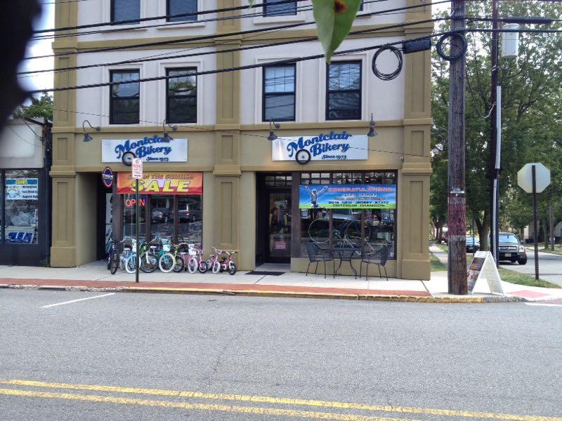 A storefront of Montclair Bakery, featuring a blue and white sign and a colorful banner advertising a summer sale. The exterior shows several bicycles lined up on the sidewalk, with additional signs visible, including an "Open" sign. The building has a classic architectural style with large windows, and street elements like a stop sign and utility pole are present in the scene.