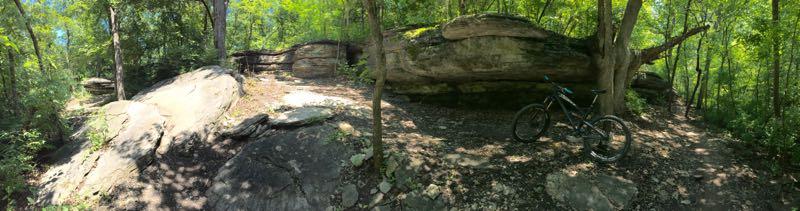 A panoramic view of a wooded area featuring large rock formations and a mountain bike resting on the ground. Sunlight filters through the green leaves of trees, creating a dappled light effect on the rocky terrain. Swope Park Trail mountain bike trail.