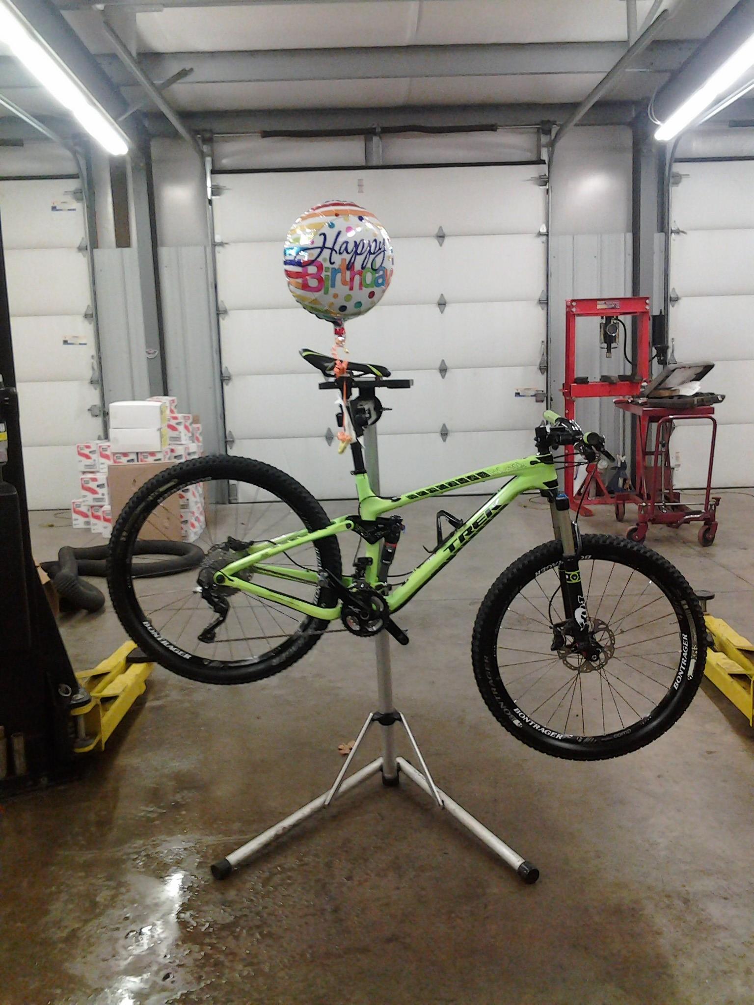 Trek Trek Fuel EX 9.8 27.5: A bright green mountain bike is displayed on a repair stand in a garage, with a "Happy Birthday" balloon attached to the seat. The background features a clean, well-lit workshop environment, including tools and a stack of boxes.