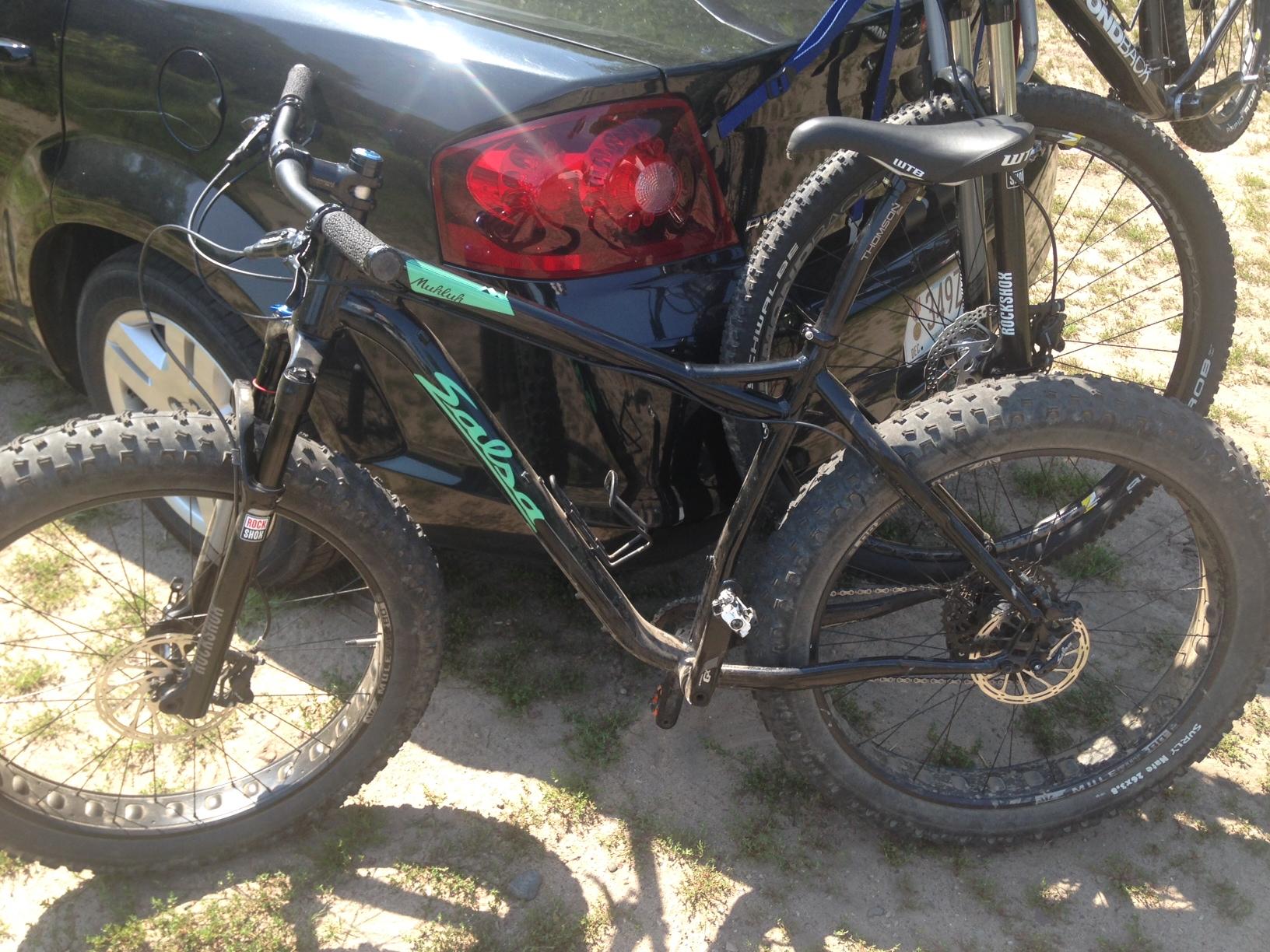 Salsa Mukluk 3: A black fat bike with green accents parked next to a black car. The bike features wide tires, a front suspension fork, and is partially obscured by another mountain bike in the background. The scene is set on a sandy area with patches of grass.