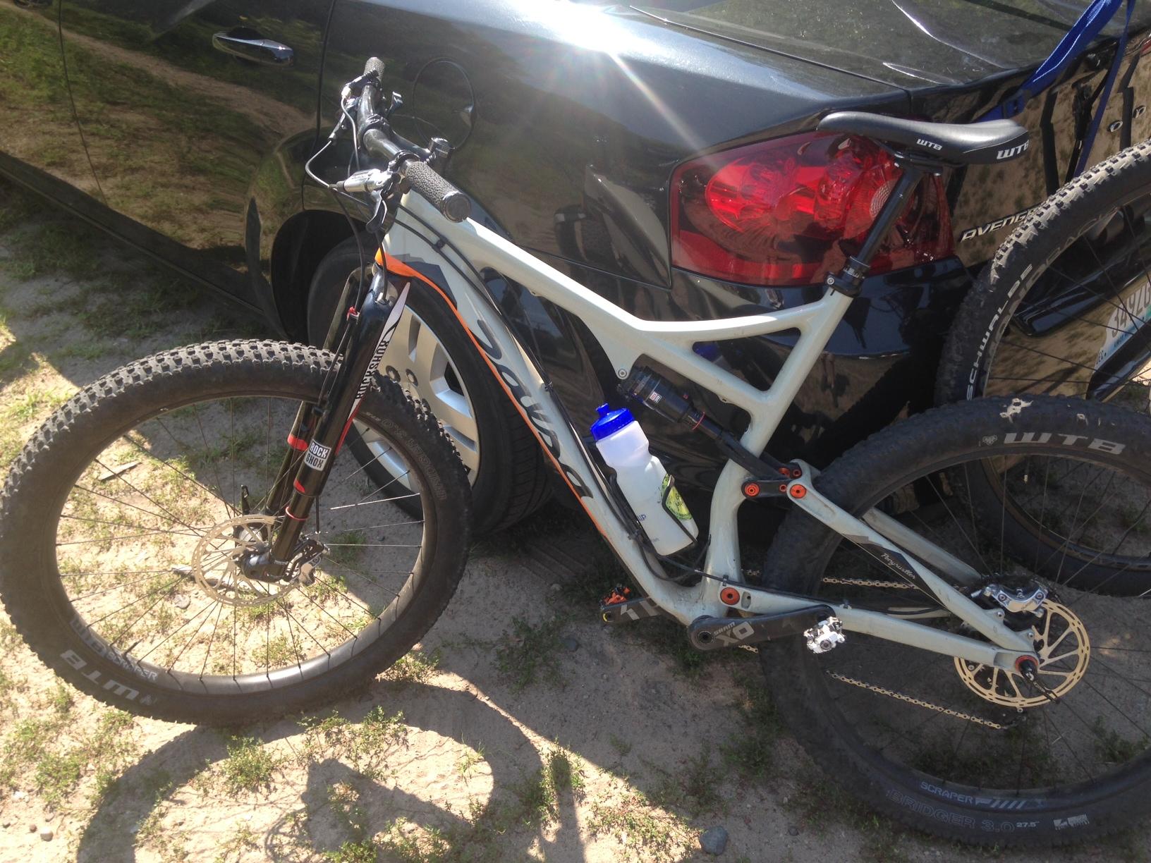 Salsa Pony Rustler Carbon XO1: A mountain bike parked next to a black car, featuring a light-colored frame with orange accents, thick tires, and a water bottle mounted on the frame. The background shows grass and sandy ground.