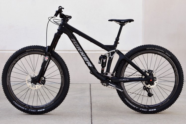 A black mountain bike with a sleek frame, featuring a suspension system and wide tires. The bike is positioned against a light-colored wall, showcasing its design elements and branding. The wheels are equipped with disc brakes and have a rugged tread pattern suitable for off-road terrain.