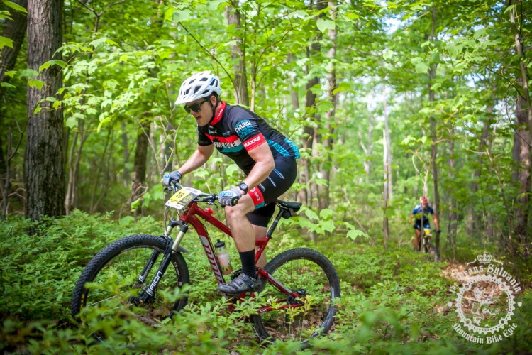 A mountain biker navigates a wooded trail, wearing a helmet, sunglasses, and a black and red cycling outfit. The bike is red with distinct tires, and the background features lush green foliage and trees. Another cyclist is visible in the background, slightly blurred, indicating movement on the trail.