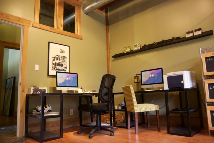 A cozy workspace featuring two desks with iMac computers, a black swivel chair, and a patterned yellow chair. The walls are painted green, accented by a wooden shelf displaying small items and a framed picture. Natural light comes in through a window above, enhancing the welcoming atmosphere of the room.