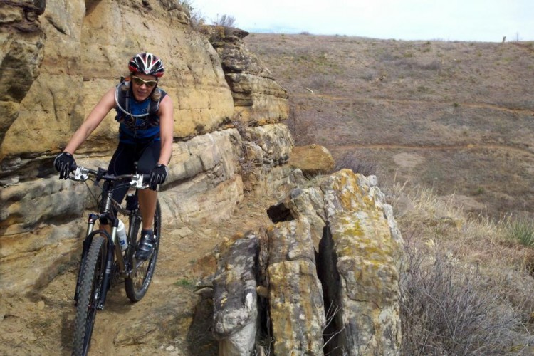 A person biking along a narrow path carved between rocky cliffs, wearing a helmet and sunglasses, with a focused expression. The terrain is rugged and arid, with sparse vegetation and a distant hillside visible in the background.