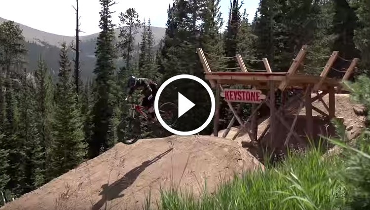 A mountain biker performs a jump off a dirt ramp, with a wooden platform labeled "Keystone" in the background, surrounded by tall trees and a mountainous landscape.