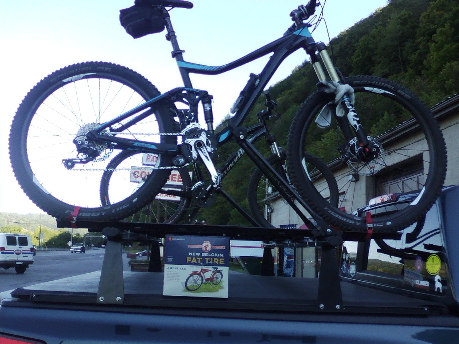 A mountain bike secured on a bike rack atop a truck, with a scenic background and a poster of New Belgium Fat Tire beer in the foreground. The bike features thick tires and a sleek frame, and the scene suggests an outdoor adventure. Allegrippis Trails mountain bike trail.